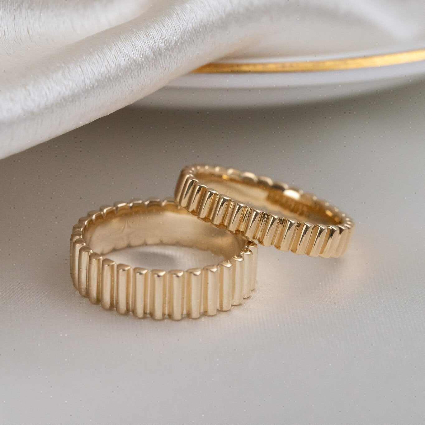 Two gold Drew bands with a ribbed texture, displayed on a white surface next to a silver ring with a similar ribbed texture.