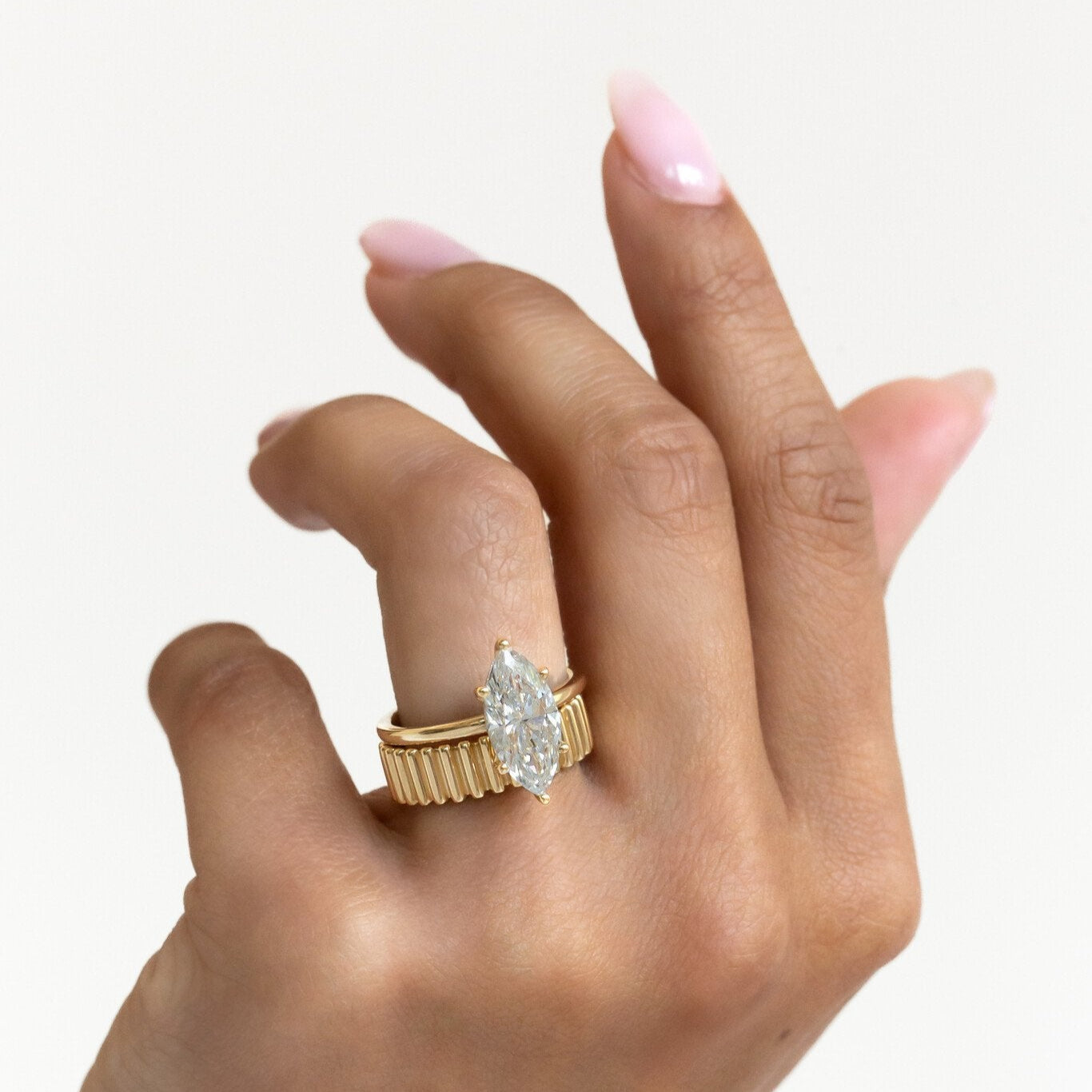 Gold Drew band with a ribbed texture stacked with a Maeve marquise diamond engagement ring on a hand against a white background.