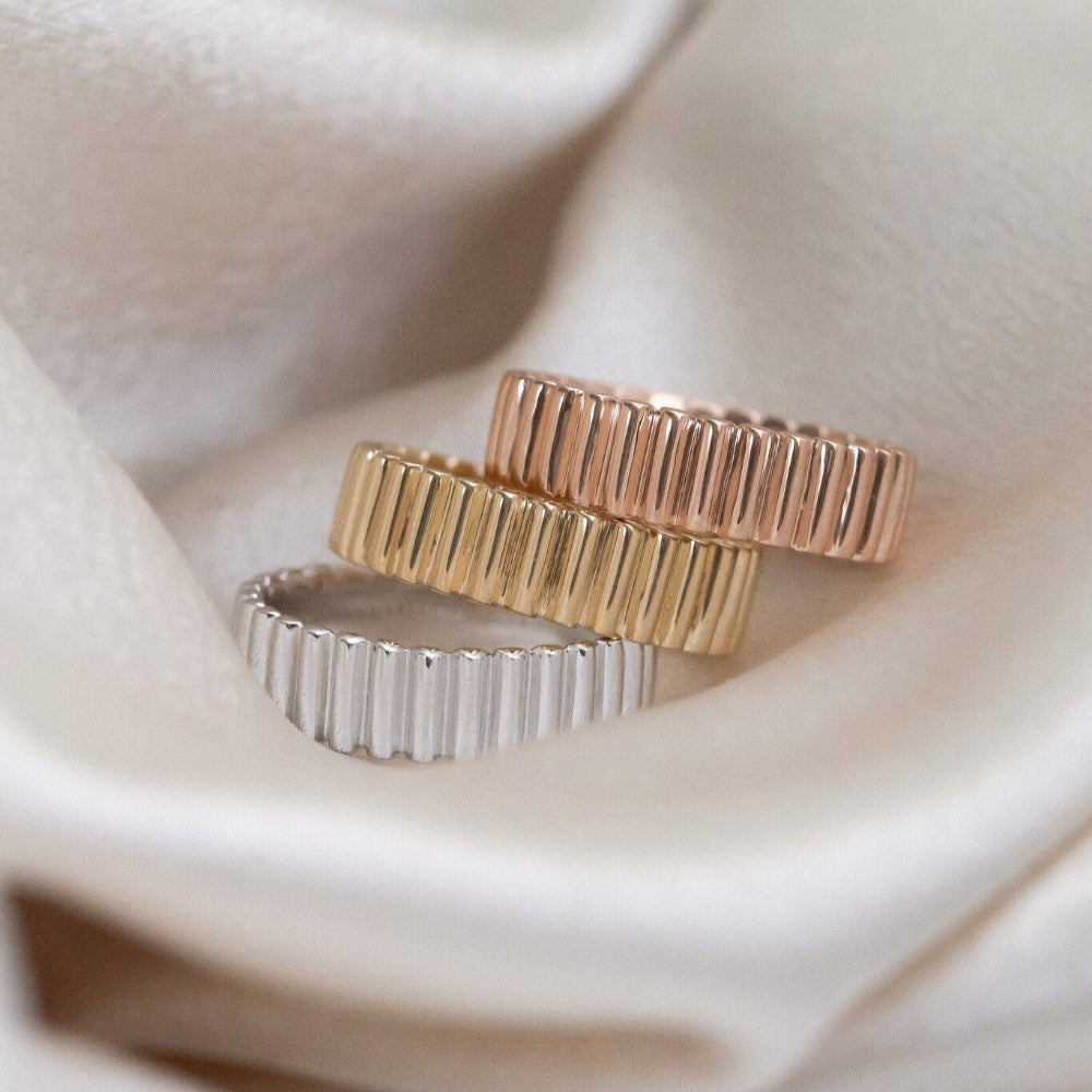 Three Drew bands with ribbed texture in white, yellow, and rose gold on a soft fabric background.
