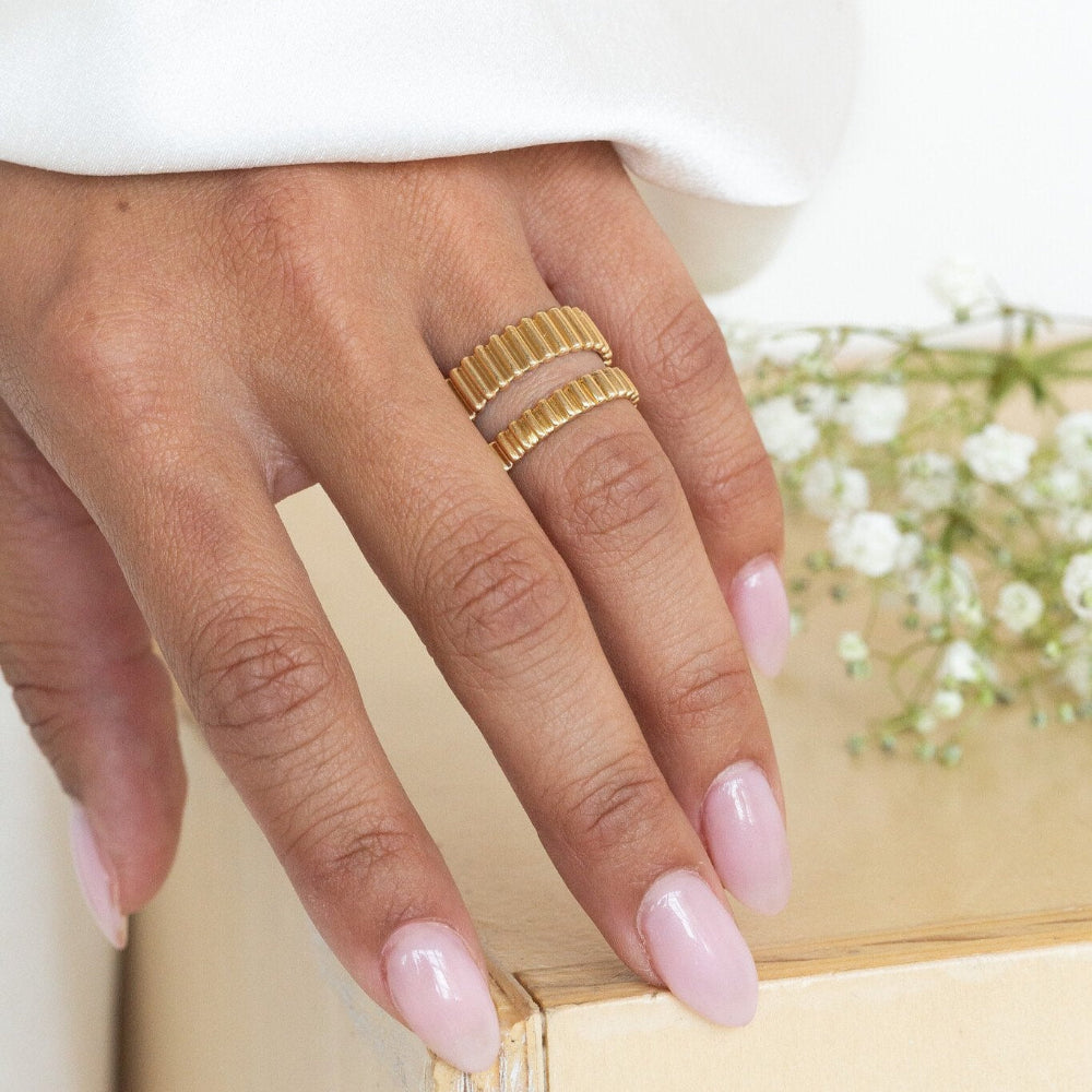 Hand wearing two gold Drew bands in different widths with a blurred background.