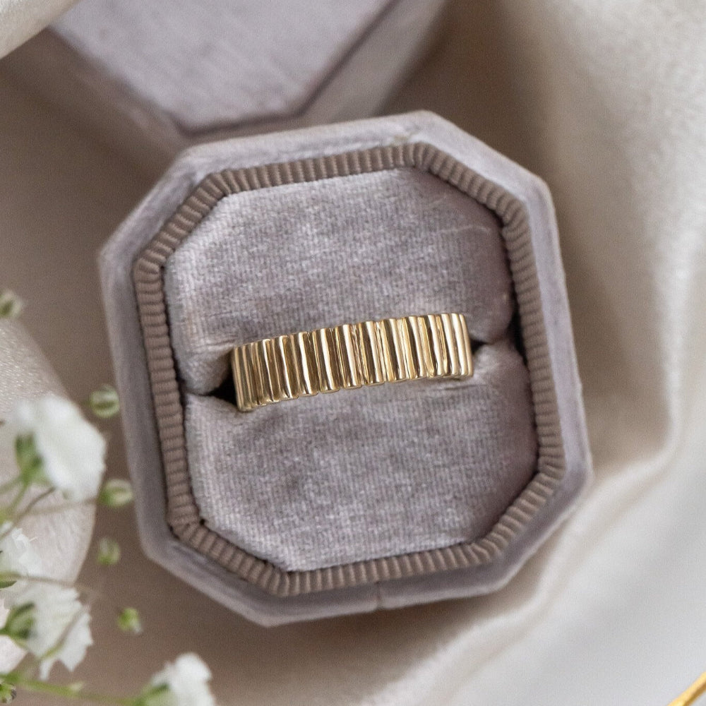 Gold Drew ribbed band in a gray velvet box on a neutral background.
