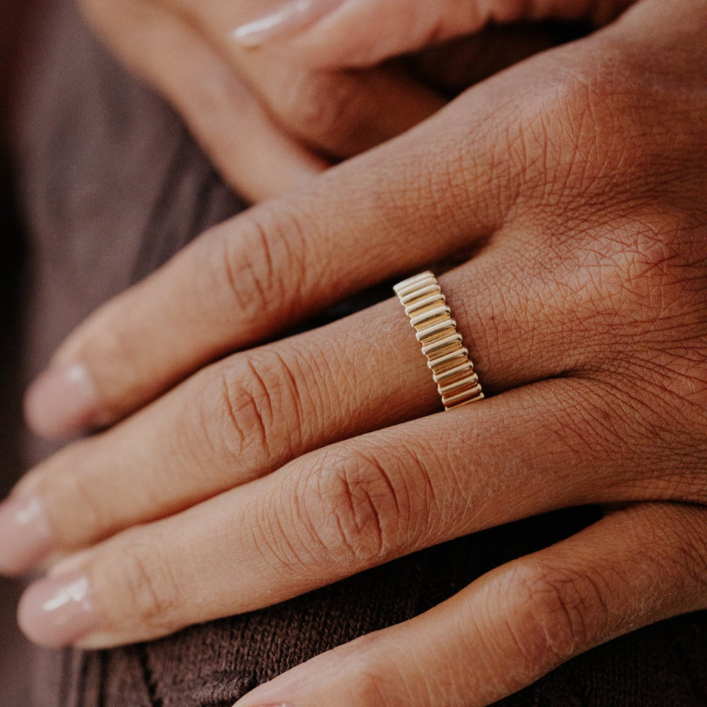 Close-up of a hand wearing a gold Drew ribbed band with a blurred background.