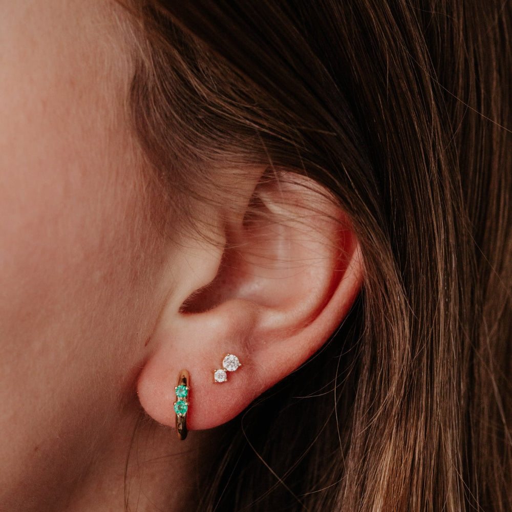 Close-up of an ear wearing two earrings: a gold one with green emerald gemstones