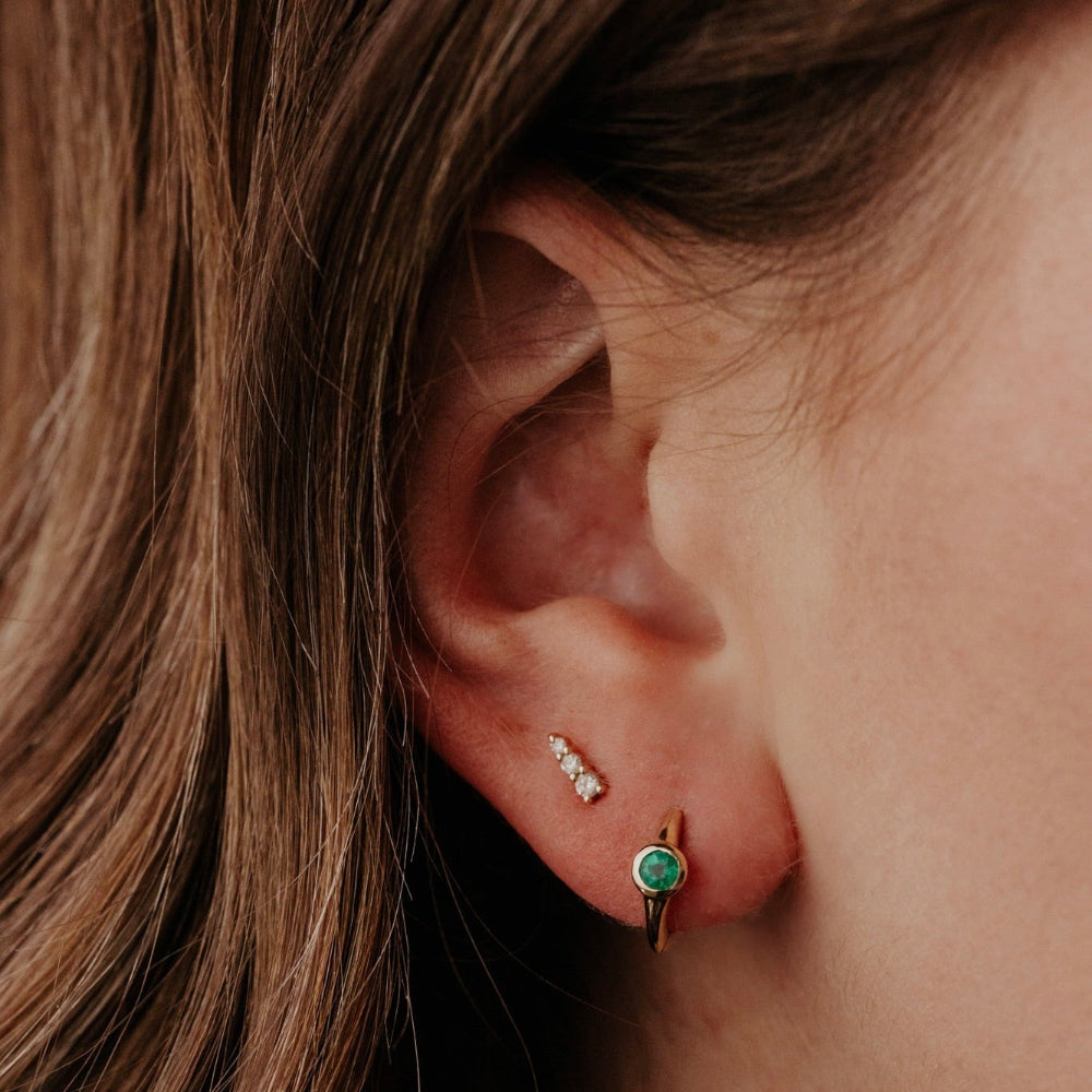 Close-up of an ear wearing gold earrings with green emerald gemstones and small diamonds.