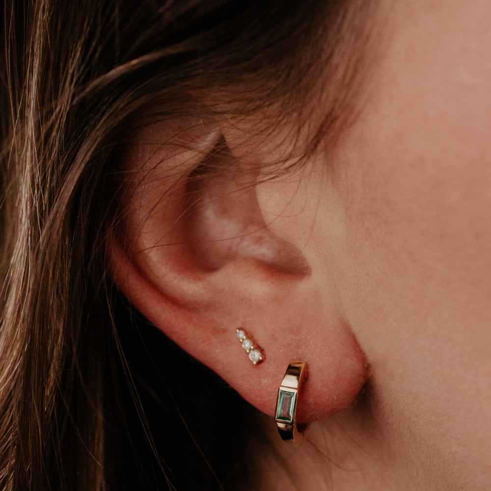 Close-up of an ear wearing gold emerald hoop earrings with a blurred background