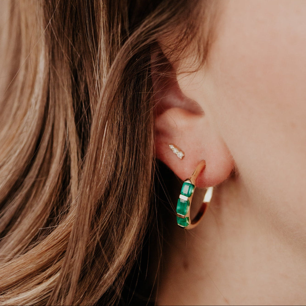 Close-up of an ear wearing a green emerald hoop earring with blurred hair and skin in the background
