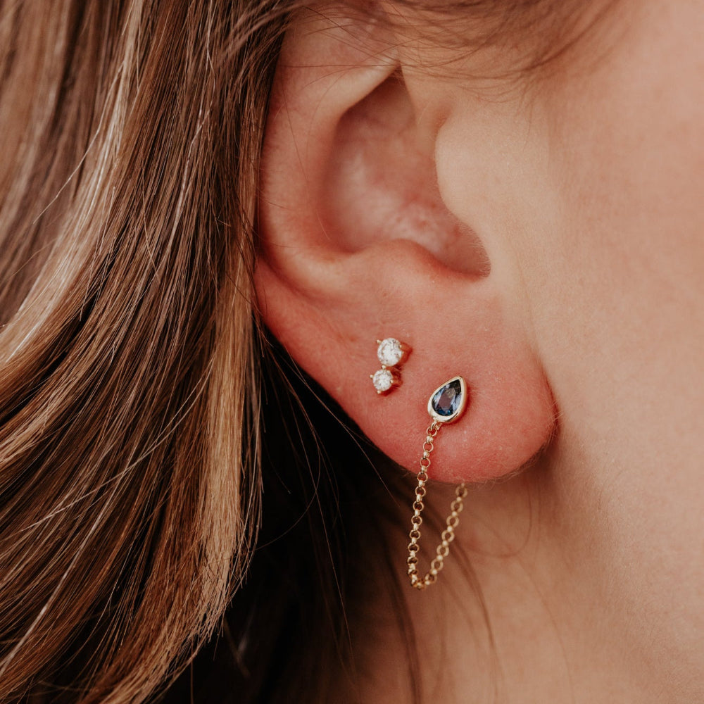 Close-up of an ear with two earrings: a small stud and a pear-shaped earring with a chain.