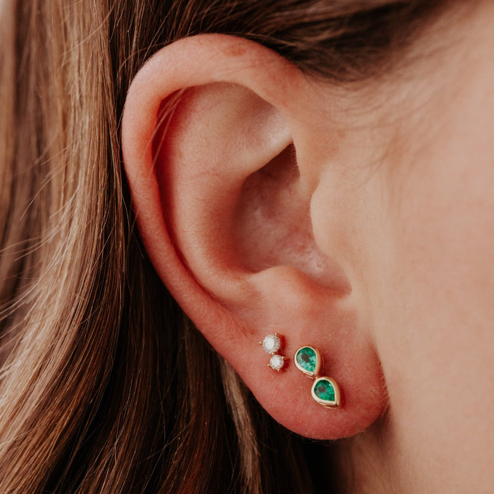 Close-up of an ear wearing two gold earrings with green gemstones and clear stones.