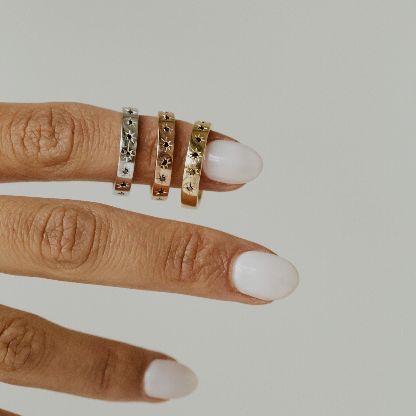 Close-up of a hand wearing three Estelle black diamond bands in white, rose, and yellow gold on a neutral background.