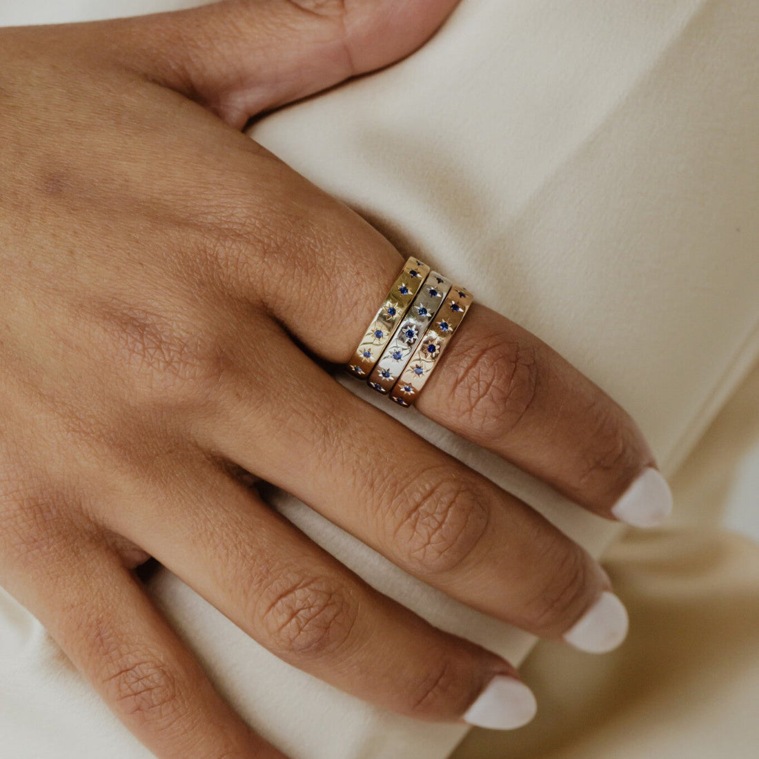 Close up of a hand wearing multiple Estelle sapphire bands in yellow, white, and rose gold on a neutral background.