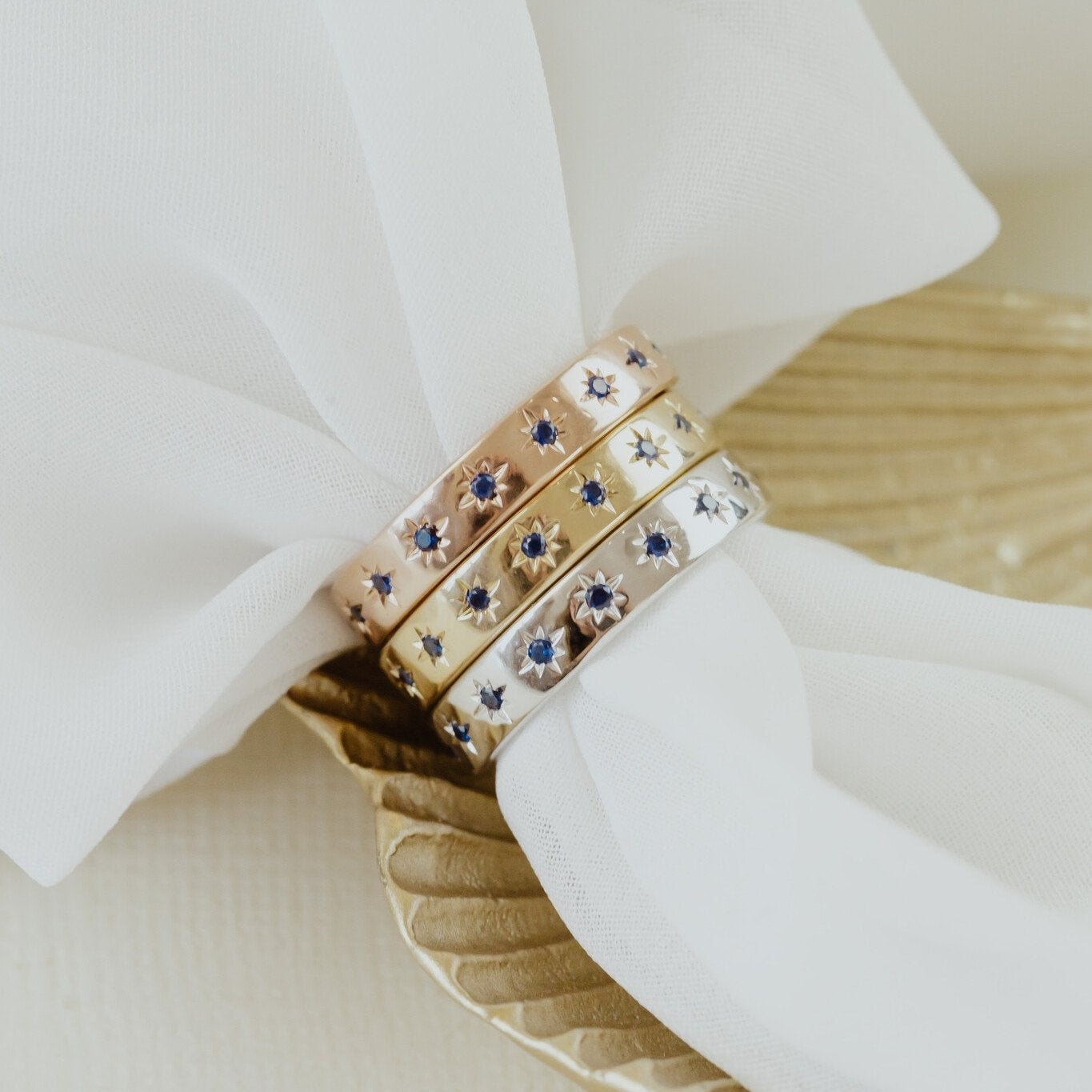 Three Estelle blue sapphire bands in rose, yellow, and white gold on a white napkin.