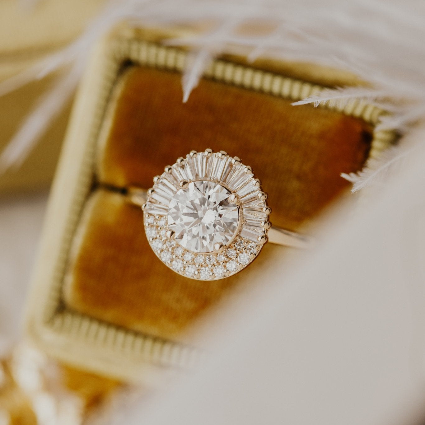 A close-up of the Florence gold engagement ring with a round center stone and a halo diamond setting, accompanied by tapered baguettes on the sides, displayed on a soft, textured surface.
