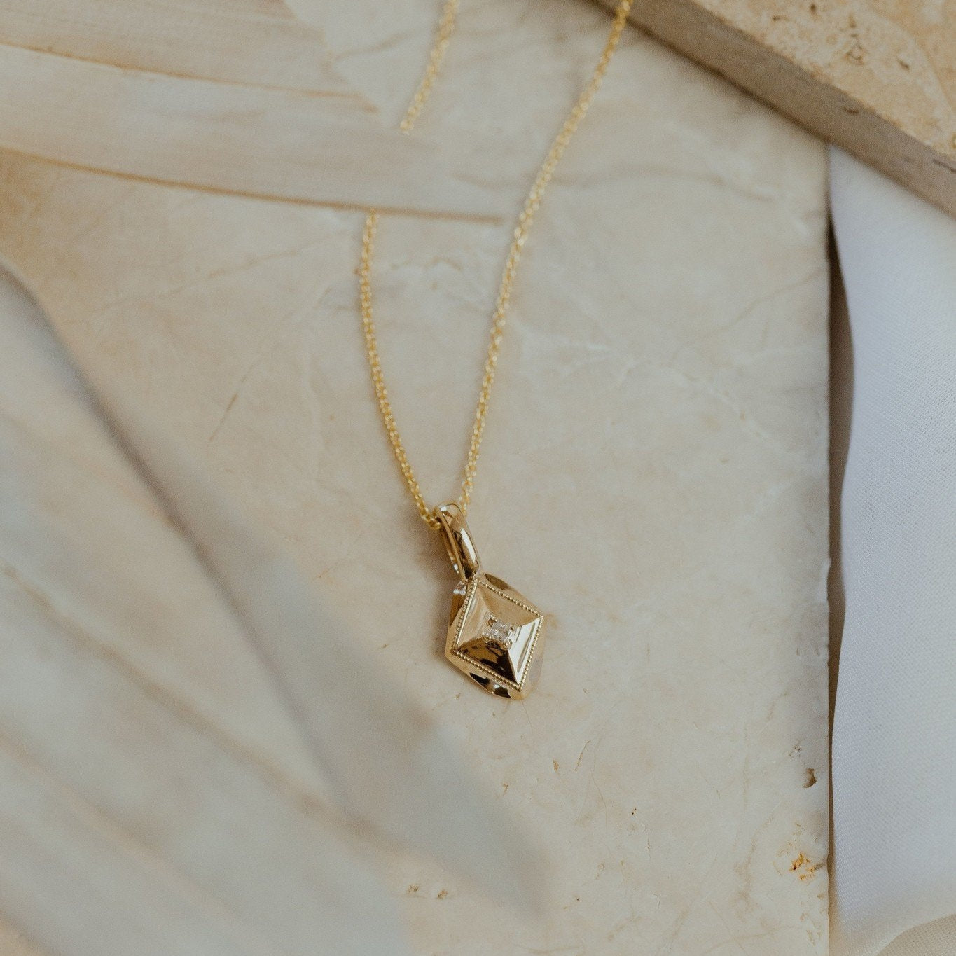 A gold necklace with a shield-shaped charm and a small diamond set in the center, displayed on a beige background.