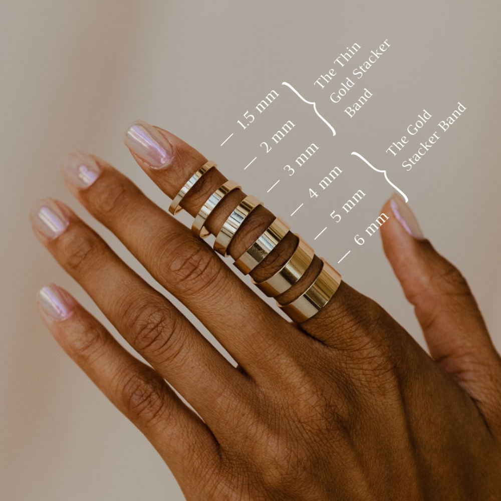 Hand wearing multiple gold stacker bands with size chart on a neutral background.