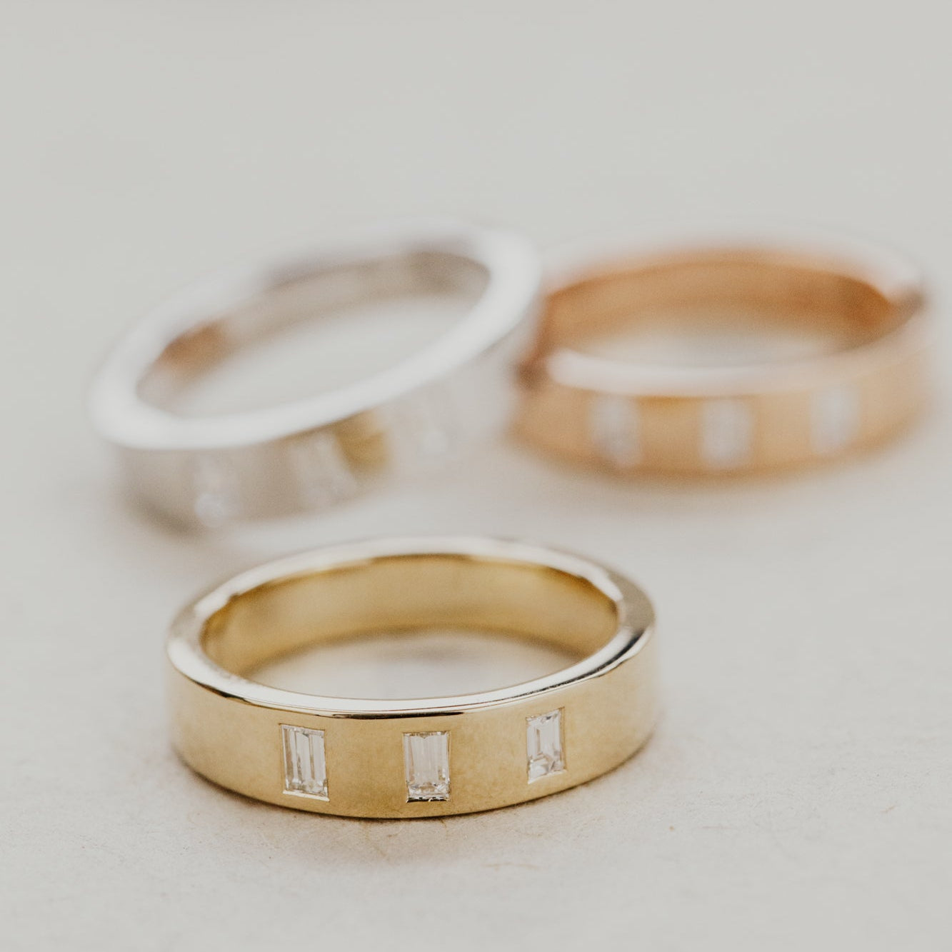 Three Jaden gold wedding bands in different metal colors with three baguette diamonds on a light background.