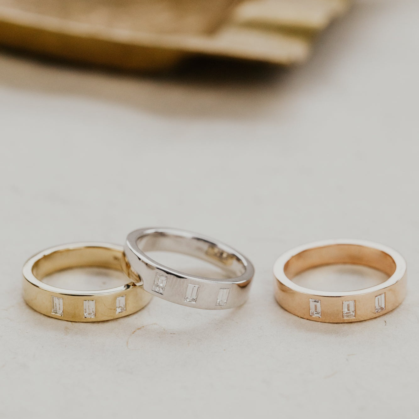 Three Jaden wedding bands in yellow, white, and rose gold with diamonds on a light surface.