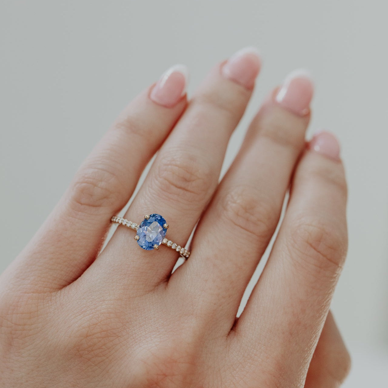 Hand wearing a Jenna engagement ring with a blue oval sapphire on a neutral background.