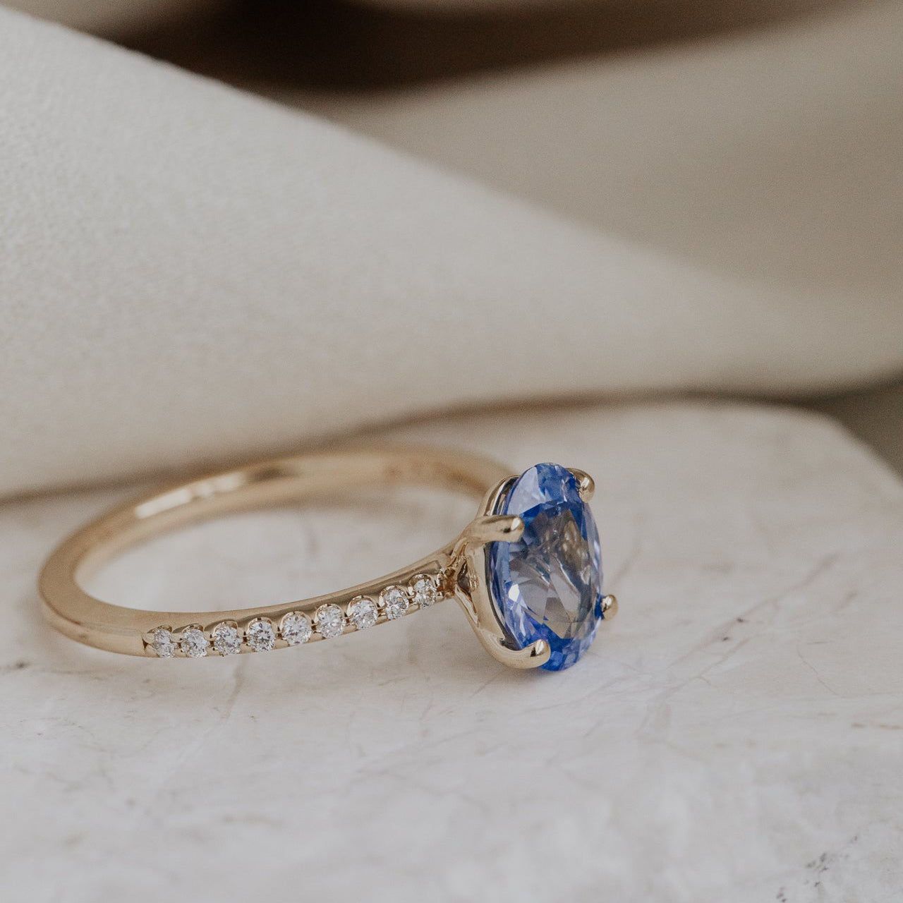Gold Jenna engagement ring with a blue sapphire and a diamond pave band on a marble surface.