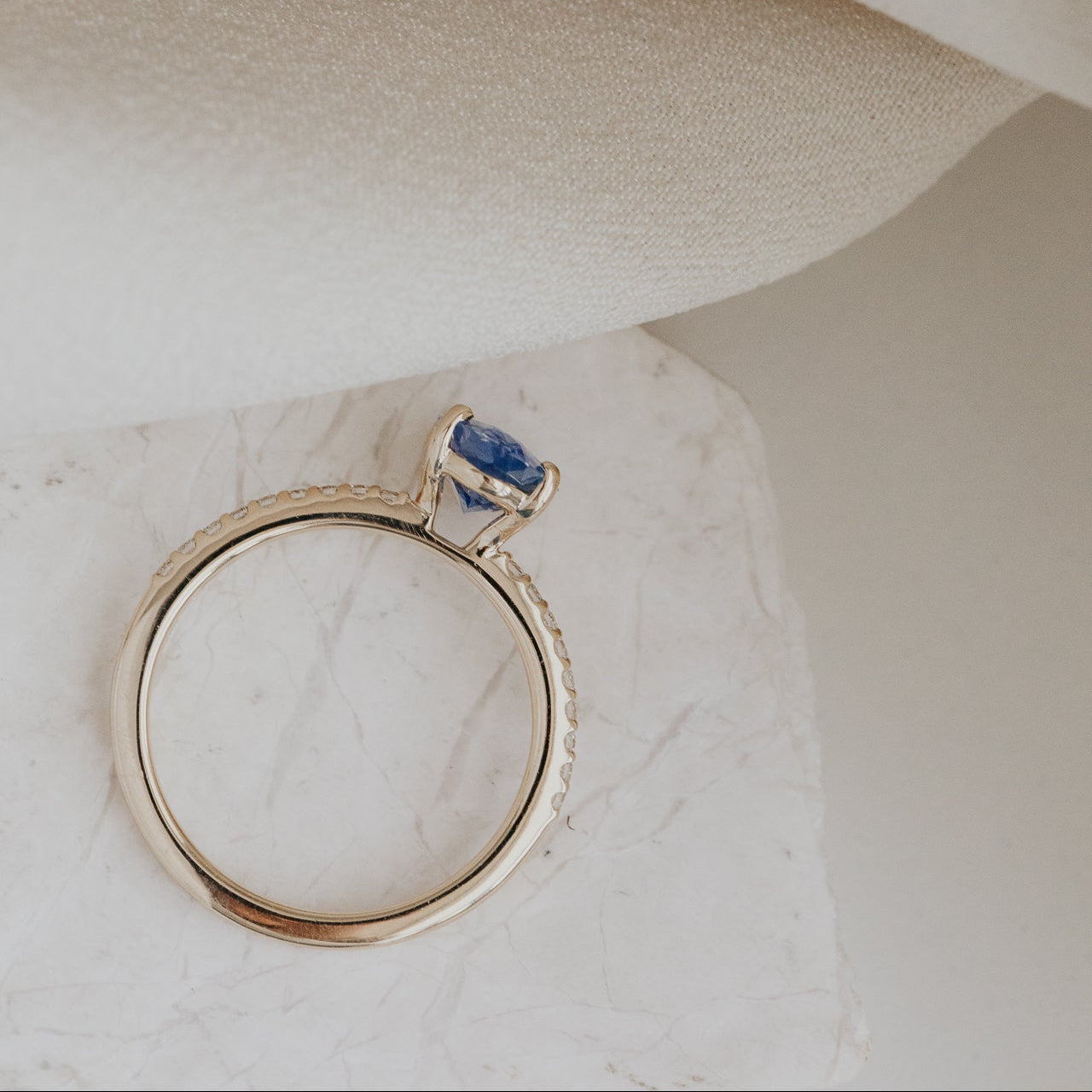 Gold Jenna engagement ring with a blue sapphire on a marble surface.