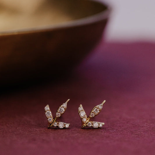 Pair of gold diamond stud earrings with leaf design on a purple surface.