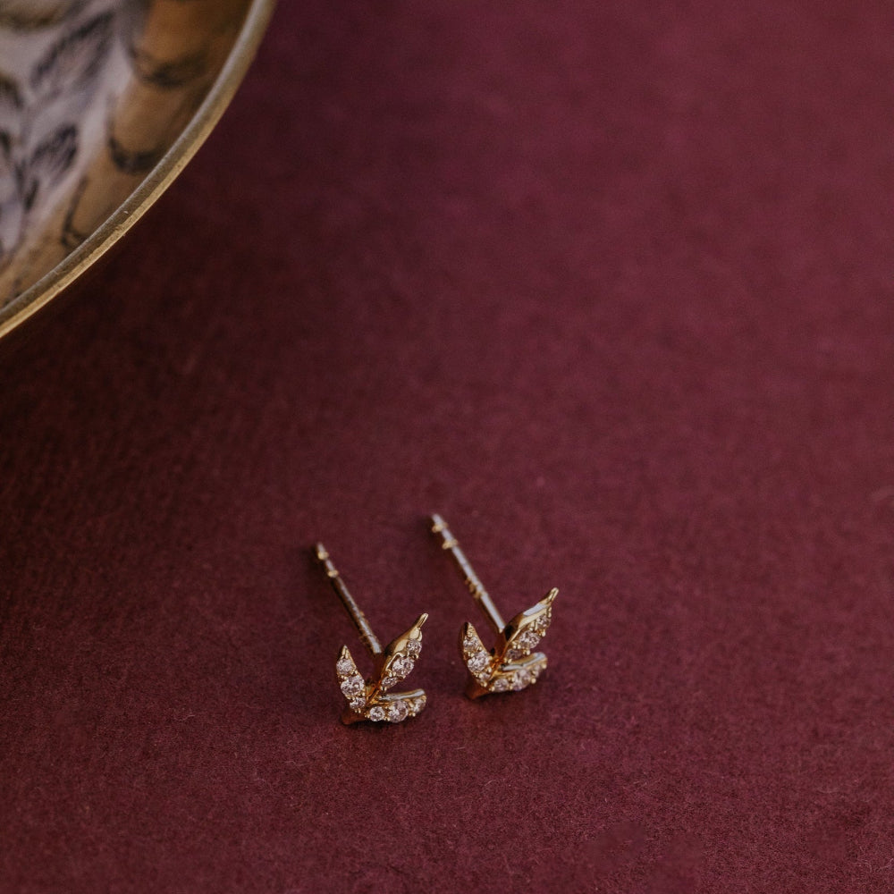 Pair of gold diamond stud earrings on a burgundy surface with a decorative object in the corner.