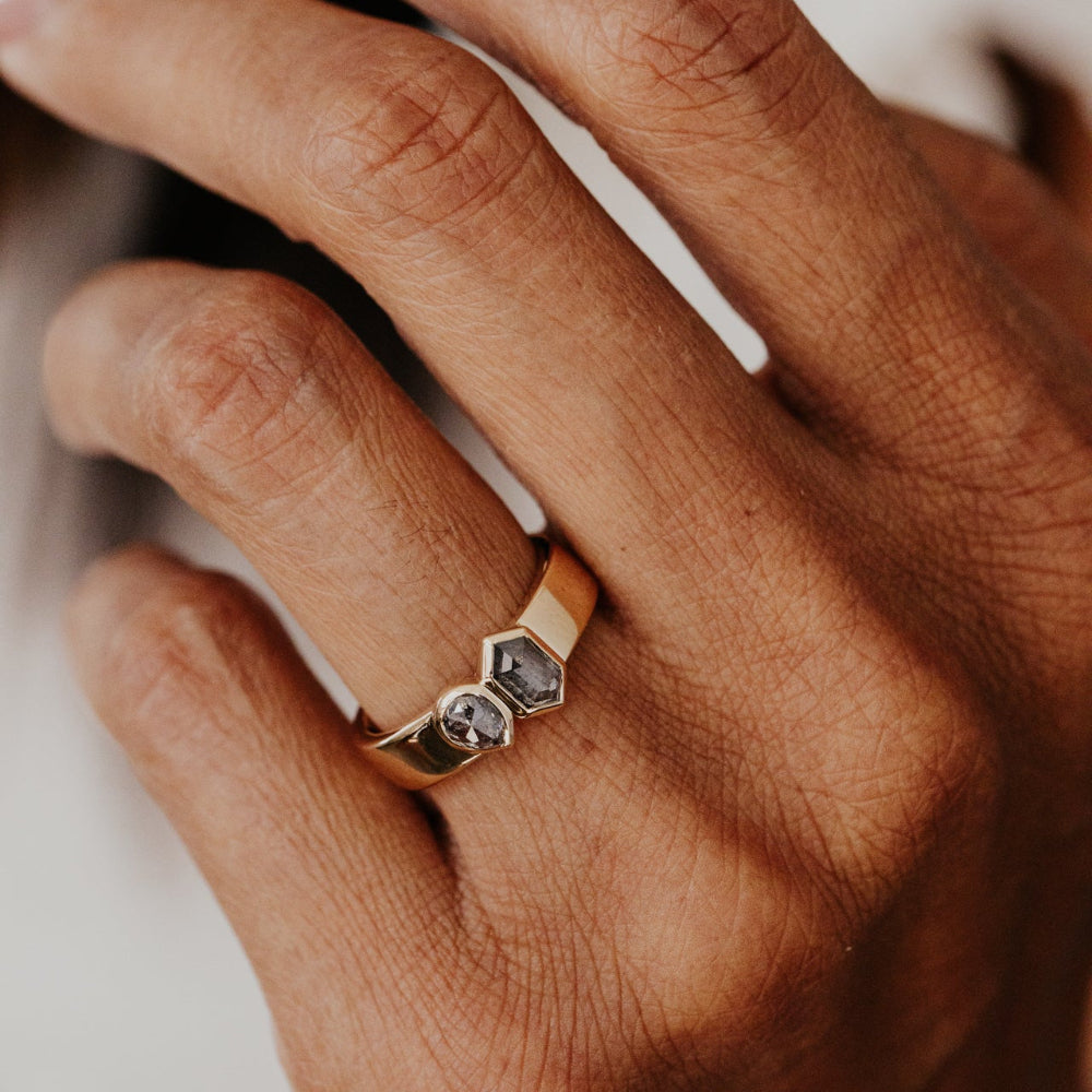 Close-up of a hand wearing a gold Madison ring with a gray hexagon and pear rustic diamond set in a bezel on a blurred background.