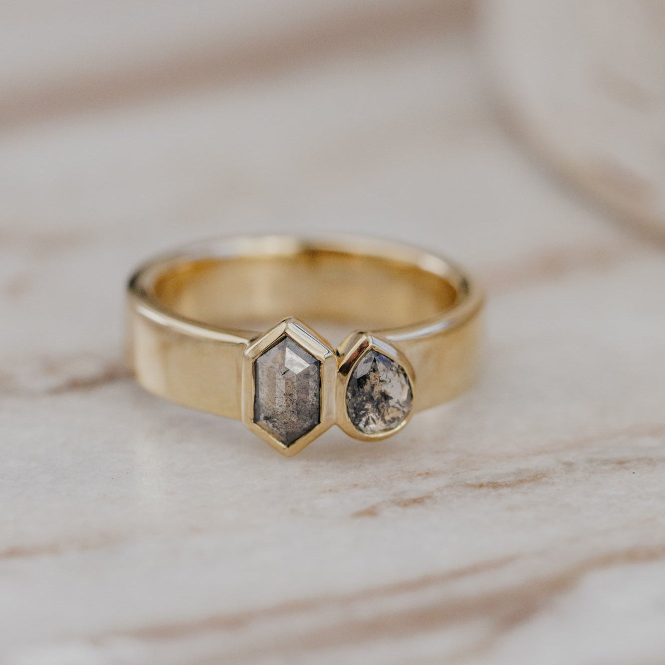 Gold Madison ring with two gray rustic diamonds set in a bezel on a marble surface.