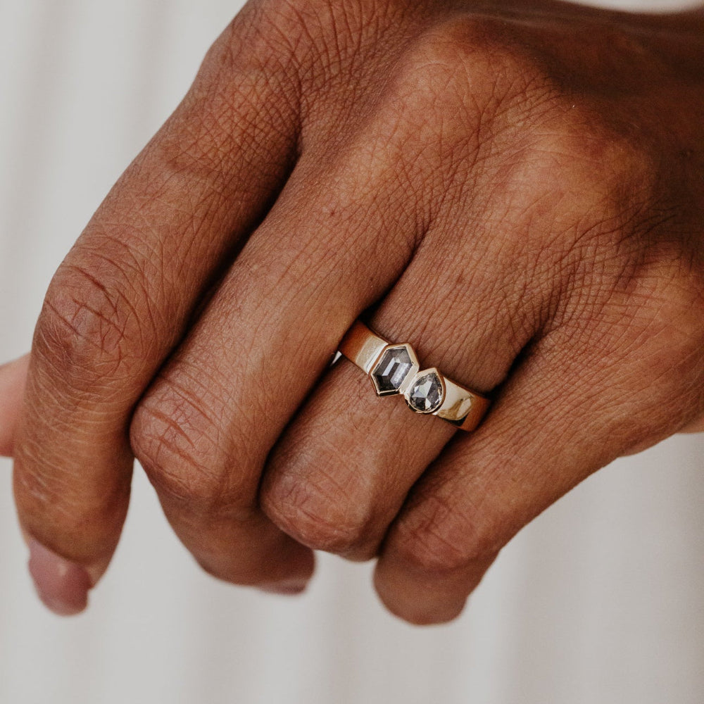 Hand wearing a gold Madison ring with two gray rustic diamonds on a neutral background.