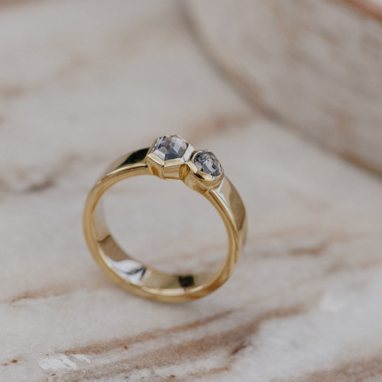 Gold Madison ring with two gray rustic diamonds on a marble surface.