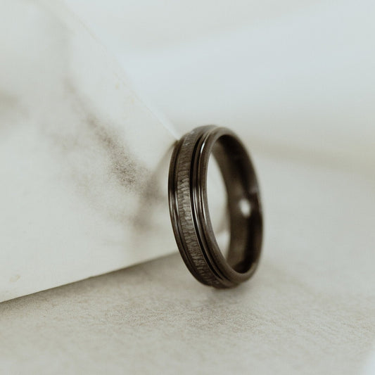 A flat band ring with a tree bark texture and grooved edges, appears to be black in color.