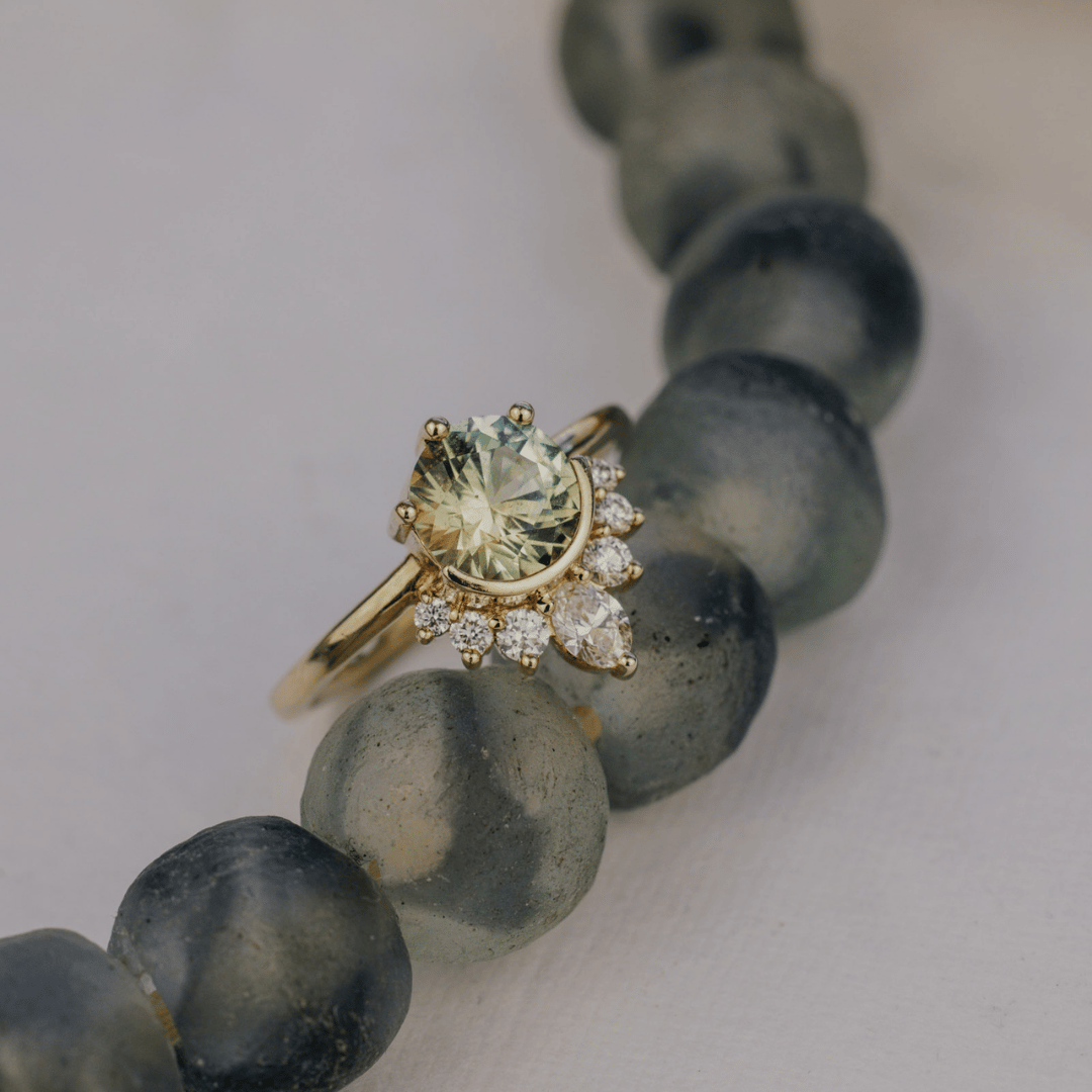 A gold ring with a round pale yellow green Montana sapphire center stone and small clear accent stones, displayed on top of a beaded necklace with dark gray translucent beads against a light background.