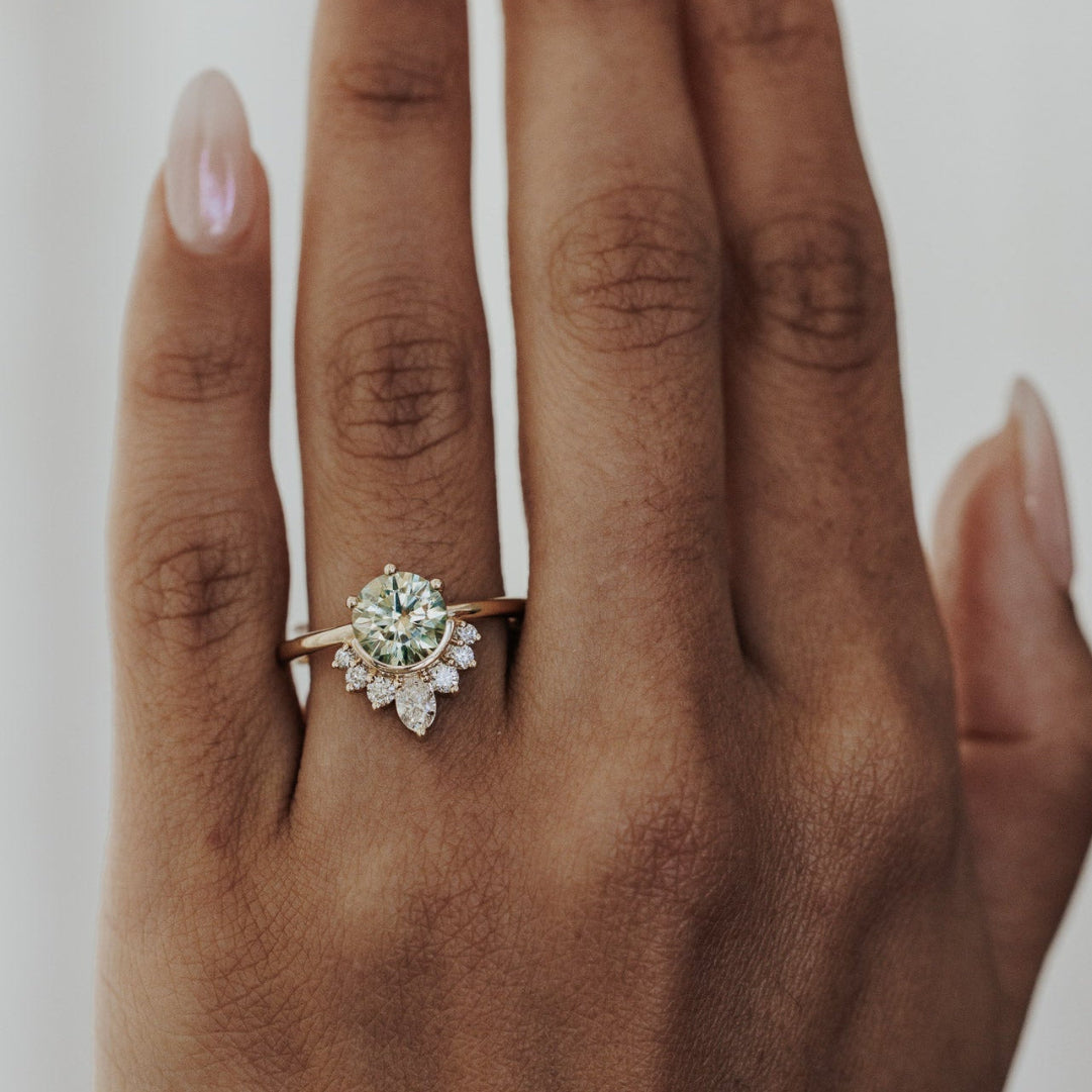 A close-up of a hand wearing a gold ring with a round-cut Montana sapphire center stone and a cluster of small clear stones below, photographed against a soft light gray background.