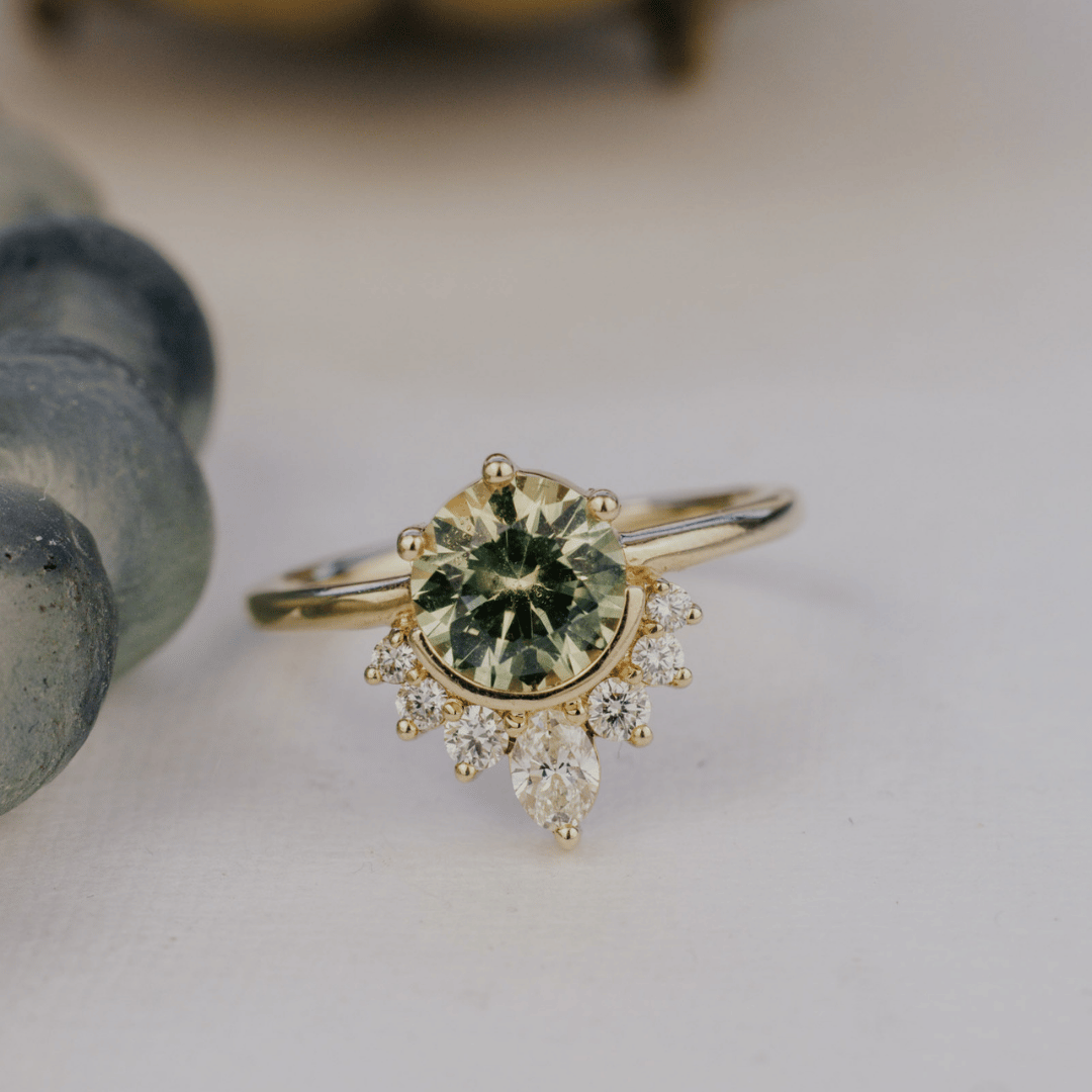 A yellow gold ring featuring a round green Montana sapphire surrounded by a halo of small clear accent stones, with a pear-shaped clear stone hanging below, photographed on a soft light background.