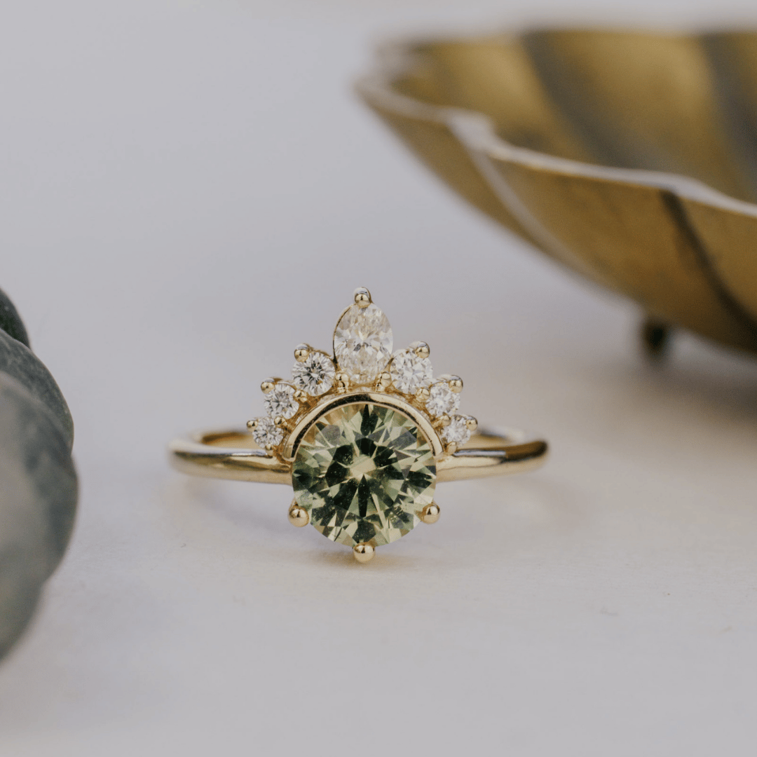 A gold ring featuring a round green Montana Sapphire surrounded by small clear stones in a sunburst setting, photographed on a light surface with soft-focus decorative props.
