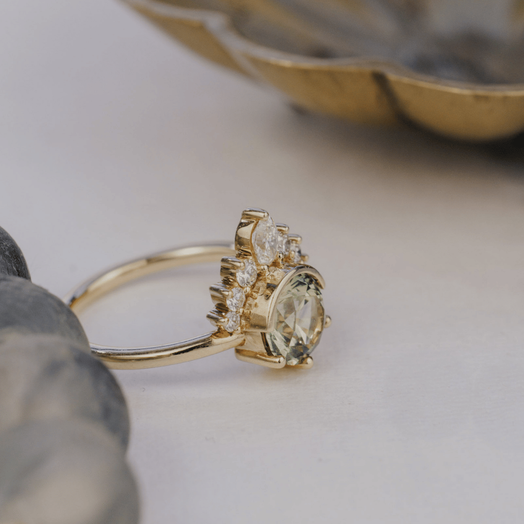 A gold ring with a large oval Montana sapphire and small accent stones, photographed on a soft light background with blurred decorative stones and a dish in the background.