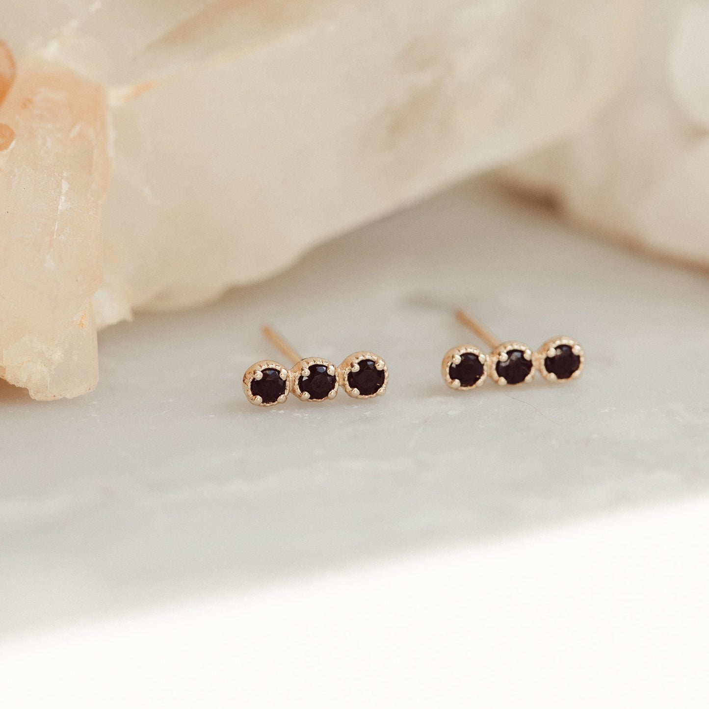 A pair of 14k gold stud earrings featuring three round black spinels set in prongs, displayed against a light-colored crystal background.