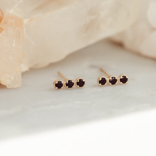 A pair of 14k gold stud earrings featuring three round black spinels set in prongs, displayed against a light-colored crystal background.