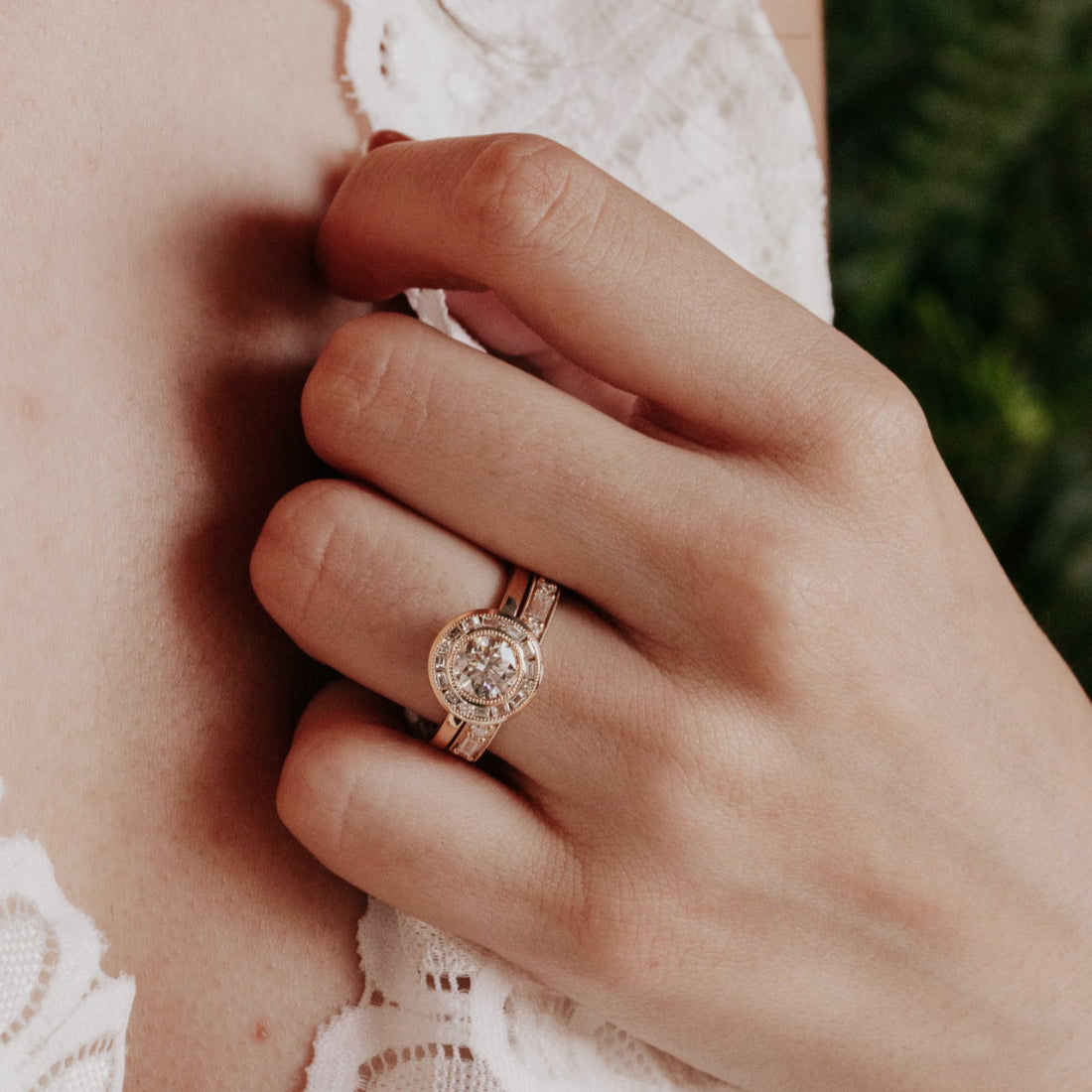 Close-up of a hand wearing a yellow gold ring with a diamond halo, against a blurred background.