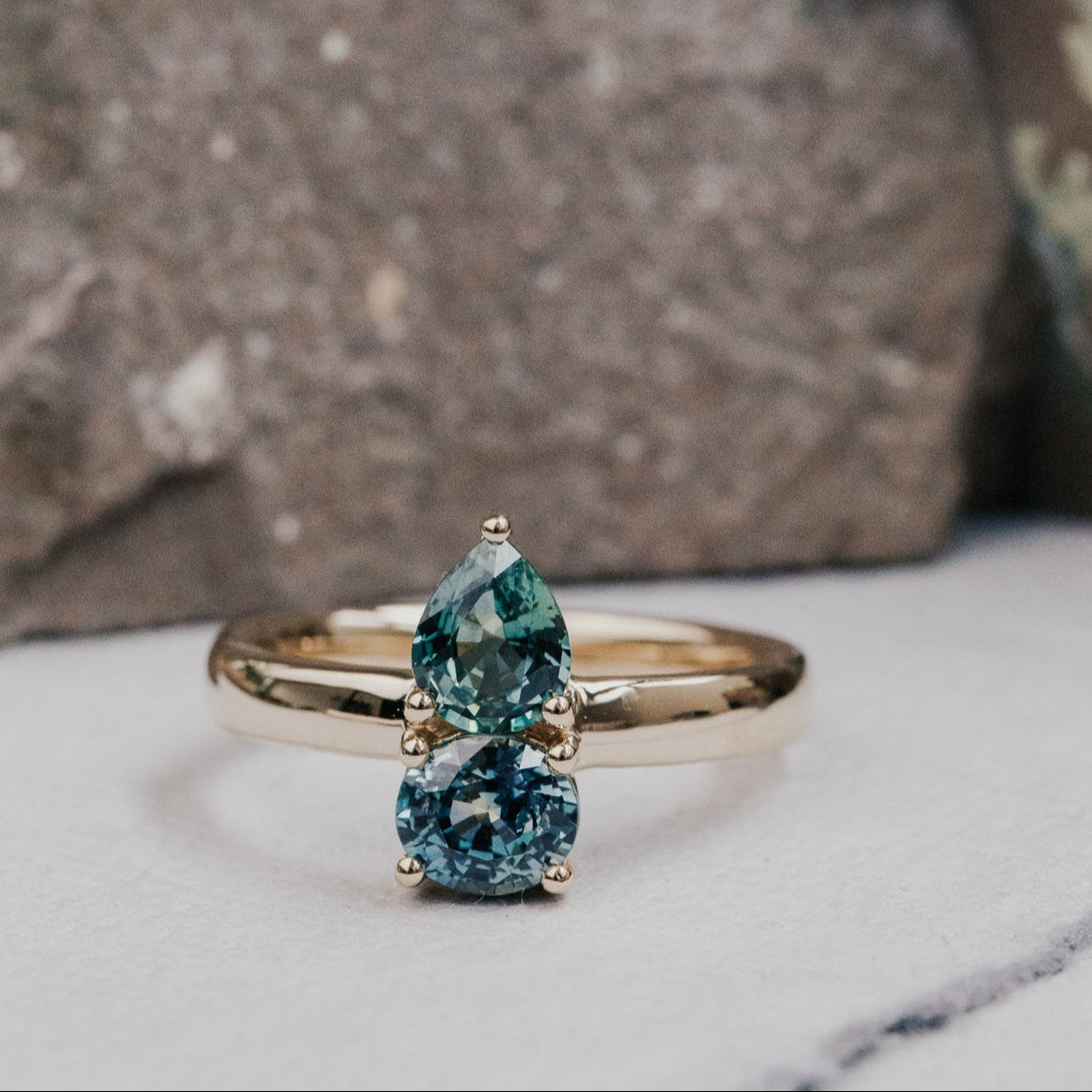 Gold ring with two green Montana sapphire gemstones on a marble surface with a stone background