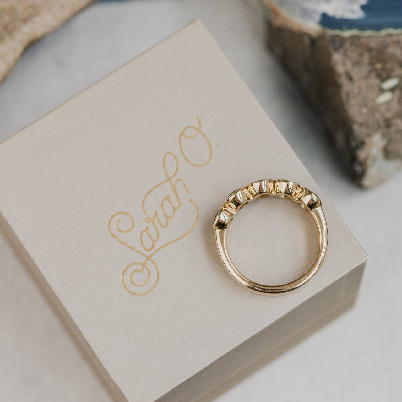 Gold ring on a box with 'Sarah' engraved, set against a natural stone background