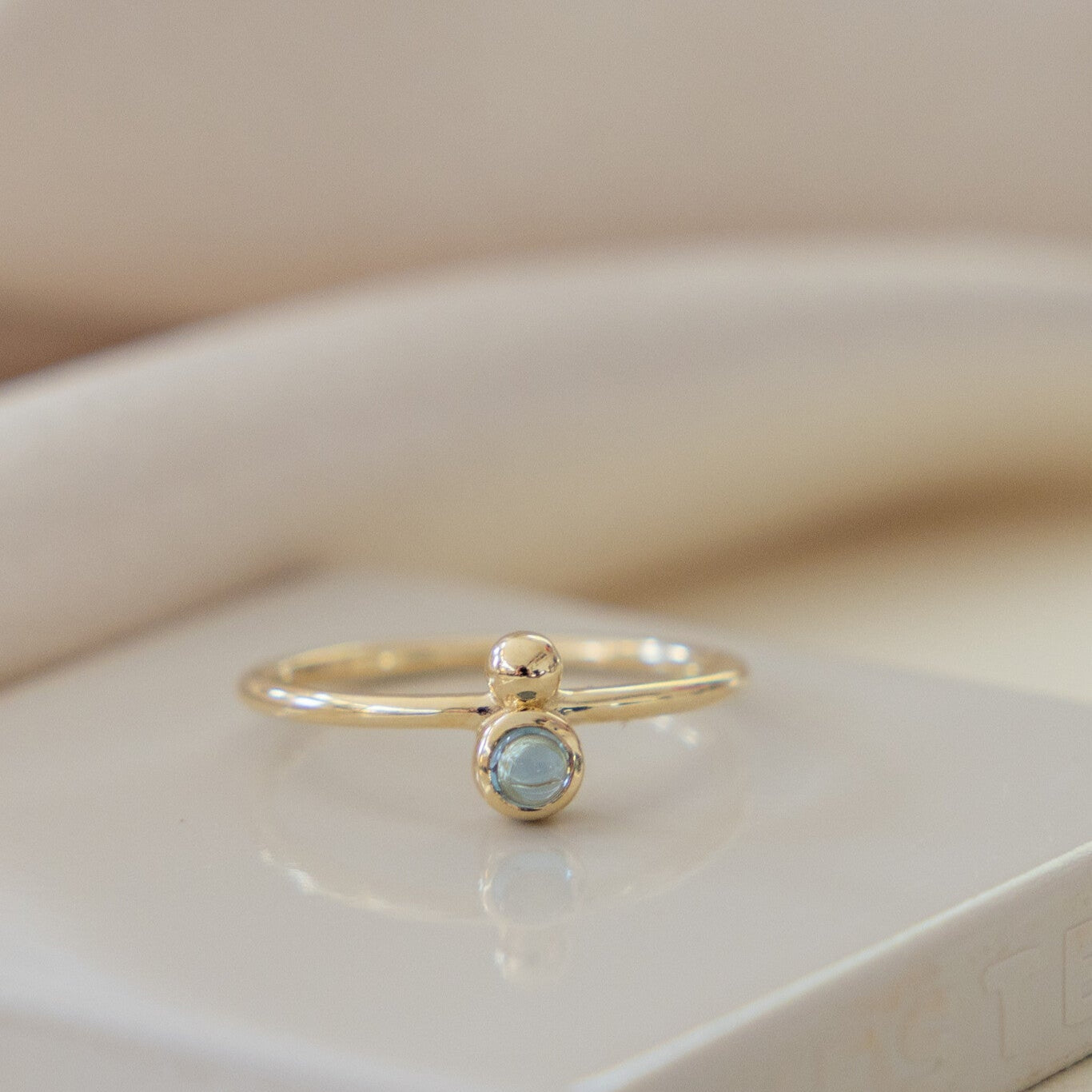A 14k yellow gold ring with a round London blue topaz, set in a high polish bezel, and a metal ball accent on a thin band, displayed on a white surface.