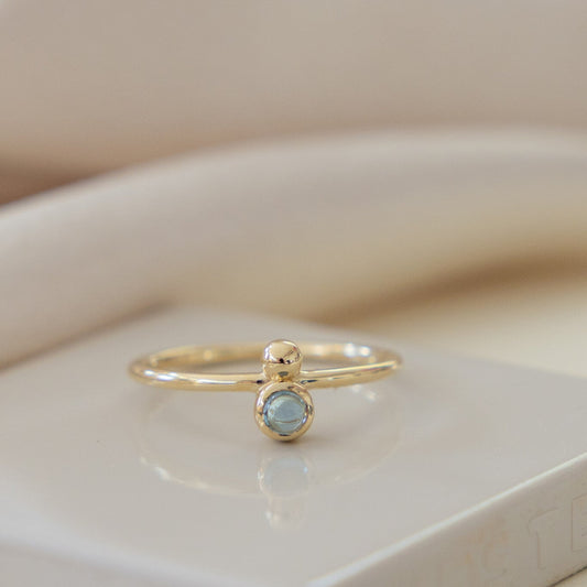 A 14k yellow gold ring with a round London blue topaz, set in a high polish bezel, and a metal ball accent on a thin band, displayed on a white surface.