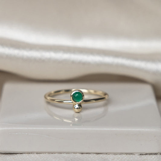A 14k gold ring with a round green agate center stone set in a high polish bezel, accompanied by a metal ball accent on a thin band, displayed on a white surface.