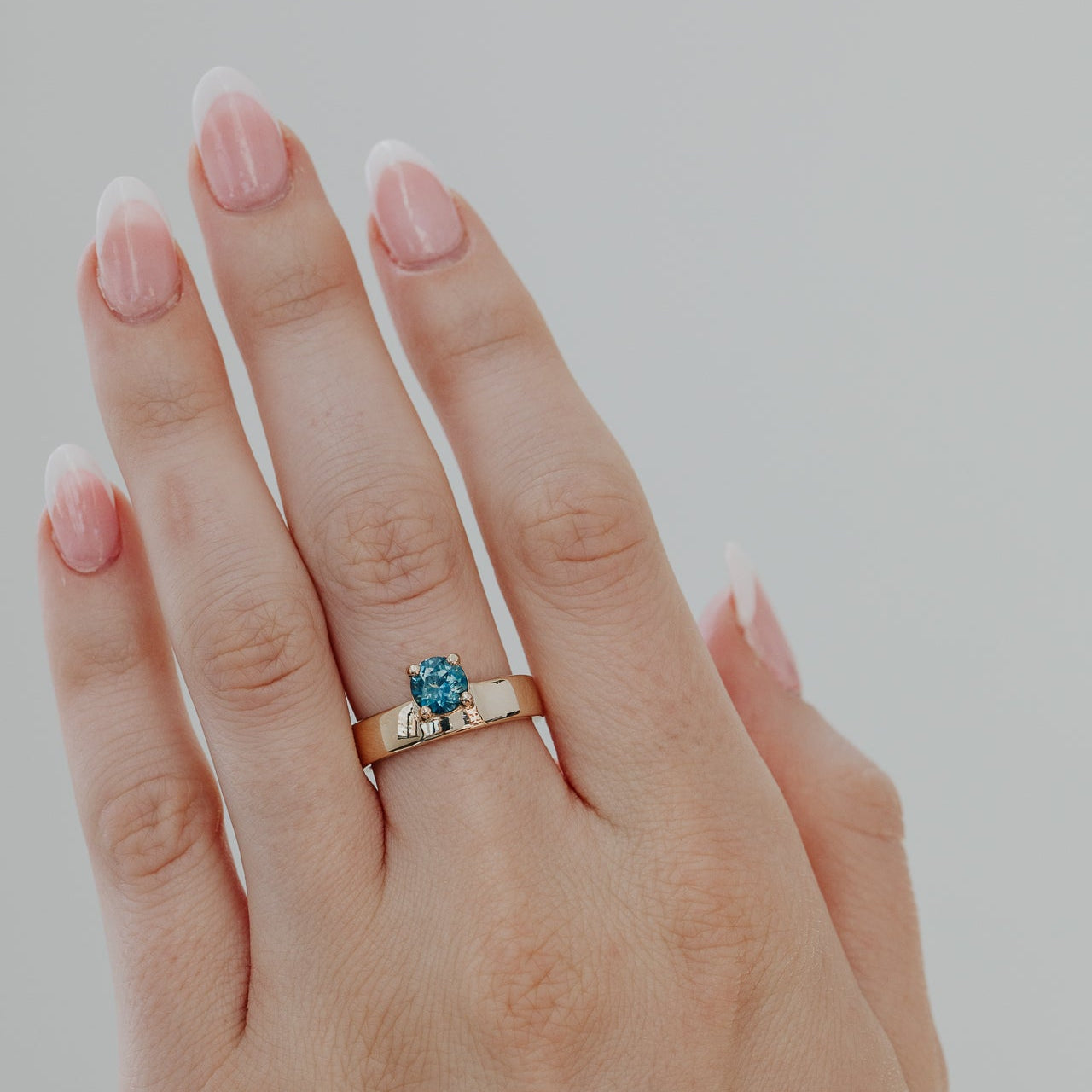 Hand wearing a gold Sezanne ring with a blue Montana sapphire on a plain background.