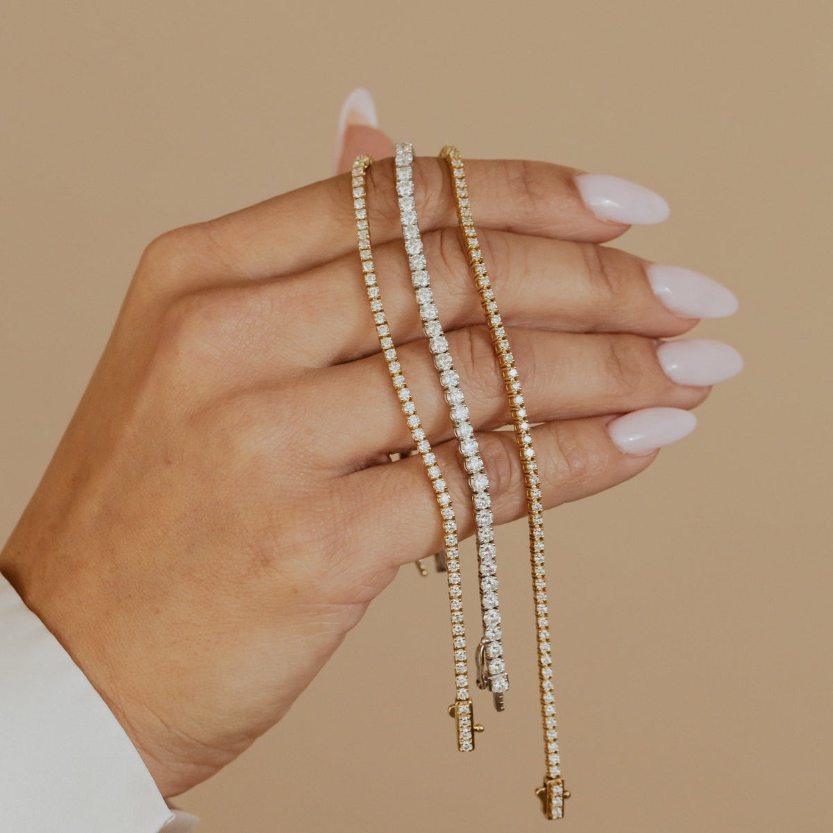 Hand holding three signature diamond tennis bracelets against a beige background