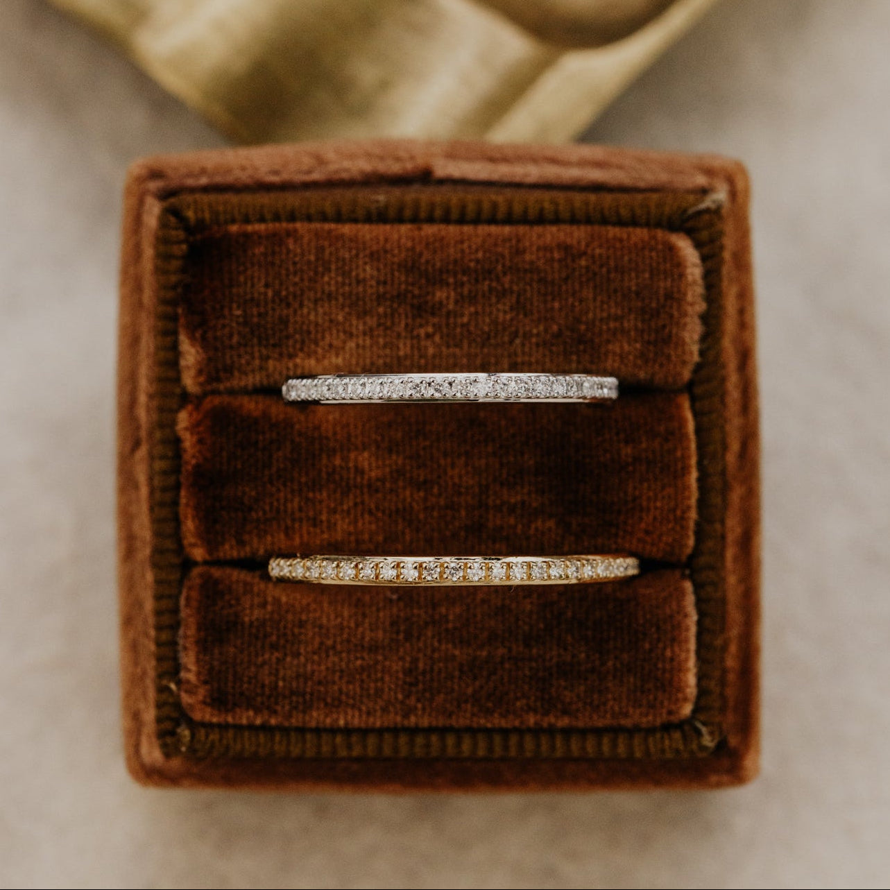 Two single row diamond wedding bands in white and yellow gold, placed in a brown velvet box on a textured surface.