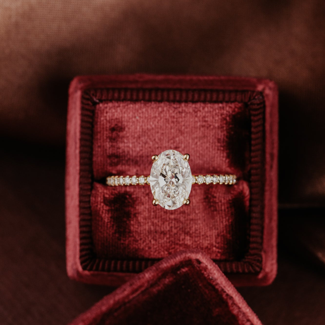 Oval shaped diamond engagement ring with a pave diamond band in a red velvet box on a brown background