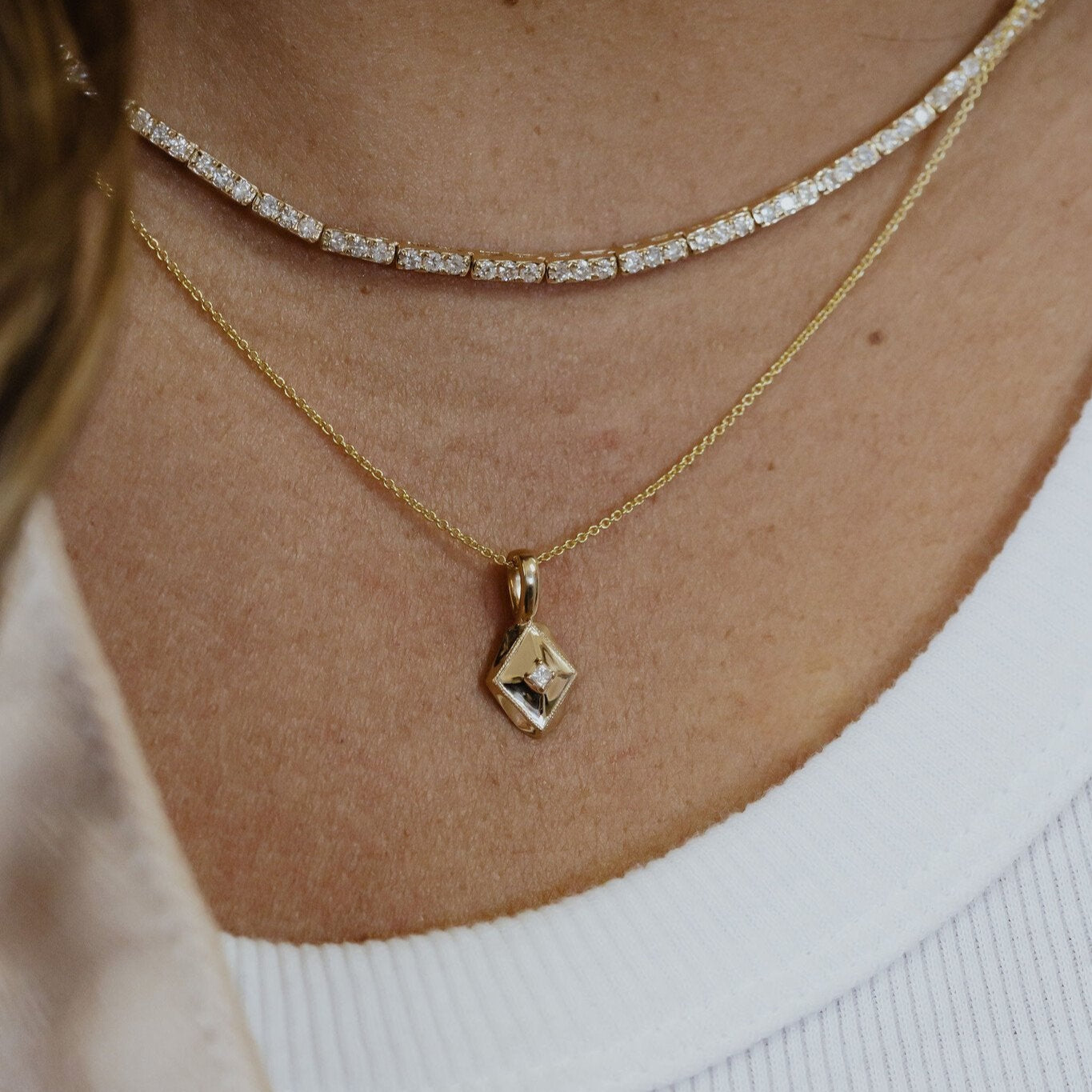 Close-up of a person wearing a diamond tennis necklace and a gold shield-shaped charm with a diamond pendant.