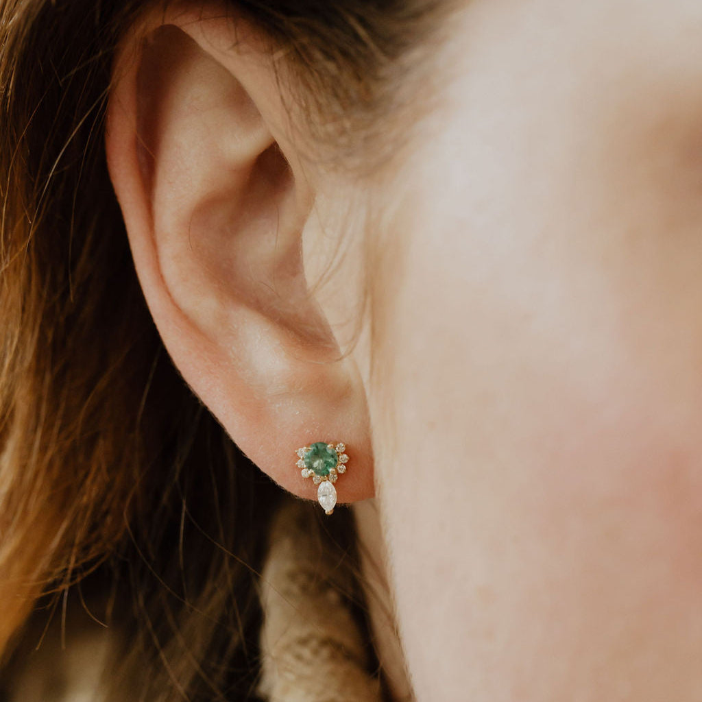 A close-up image of a person wearing 14k yellow gold stud earrings with round emeralds and a half halo design of marquise and round diamonds.