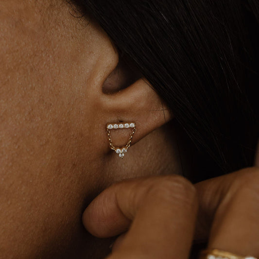 A close-up image of a person wearing 14k gold stud earrings with a pave diamond bar and three diamond dangle clusters.